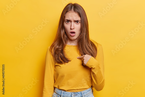 Young woman points finger at chest verbally defending herself with perplexed and puzzled expression with a challenging gaze and indignant tone exclaims who me dressed casually isolated on yellow wall photo