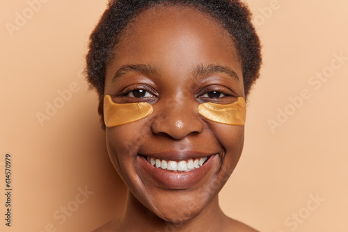 Close up shot of young African woman radiates joy as tends to her skin during beauty day at home applying golden patches under eyes for rejuvenation smiles broadly isolated over brown background