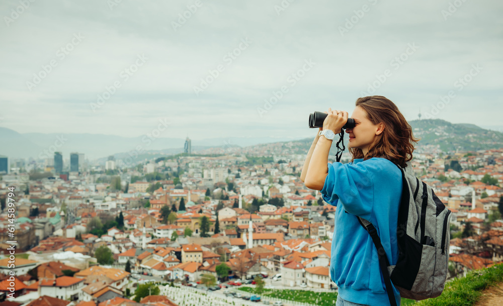 A young adult smiling female traveler looks through binoculars at a European city with orange roofs in the mountains. Travel to the Balkans, Sarajevo, Bosnia A young adult smiling female traveler looks through binoculars at a European city with orange roofs in the mountains. Travel to the Balkans, Sarajevo, Bosnia