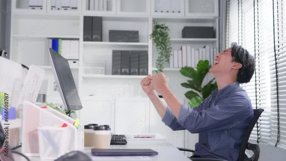 Happy young Asian businessman celebrate his success. Excited man with arms raised shouting sitting at desk in office Happy young Asian businessman celebrate his success. Excited man with arms raised shouting sitting at desk in office