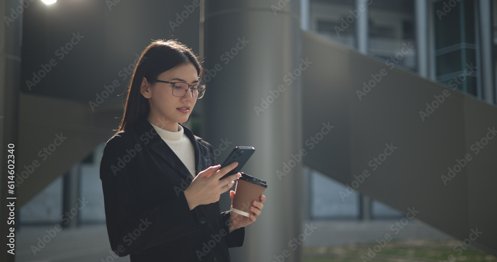 Fototapeta premium Young Asian business woman in suit holding mobile phone with takeaway coffee