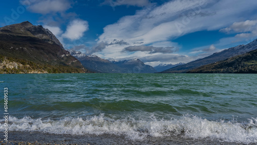 Emerald lake is surrounded by picturesque mountains. The waves are foaming and splashing on the shore pebbles. Clouds in the blue sky. Argentina. Lago Roca. Tierra del Fuego National Park.