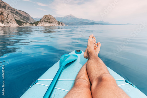 Fototapeta Naklejka Na Ścianę i Meble -  Men's legs and paddle on a supboard against the backdrop of a picturesque seascape with a rocks. Relaxation and fitness on the water