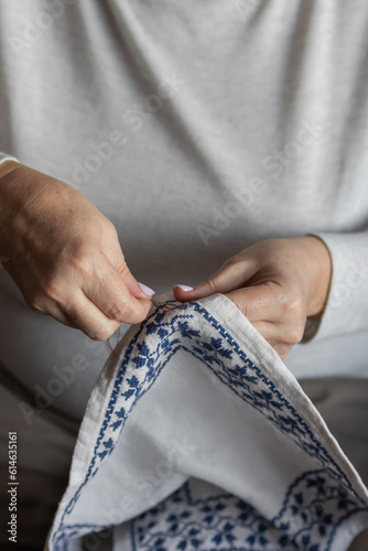 Senior woman's hands embroidering a cross-stitch floral pattern on linen fabric with needle and blue thread. Needlecraft