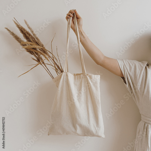 Young beautiful woman in neutral beige creamy linen dress holding sandy shopper bag with dried pampas grass over white wall. Mock up with blank copy space. Shopping