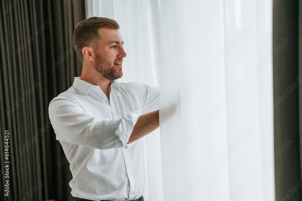 Looking through the window, holding curtains. Businessman is indoors in the hotel room Looking through the window, holding curtains. Businessman is indoors in the hotel room