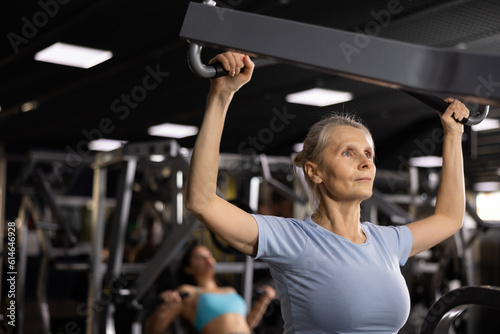 Focused aged woman working out back muscles in modern gym, performing lat pull-down on lever machine. Concept of physical activity of elderly..