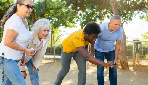 Positive multiracial mature people playing petanque game while enjoying sunshine weather outdoors in autumn