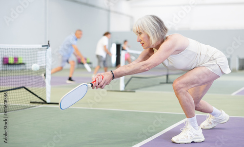 Elderly active woman bouncing ball in pickleball game on court with racket
