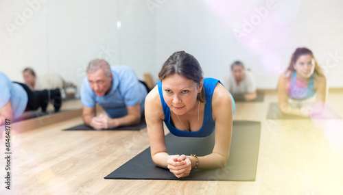 Slim middle-aged woman engaging in pilates training on mat in gym room during workout session. Persons practicing pilates in fitness studio