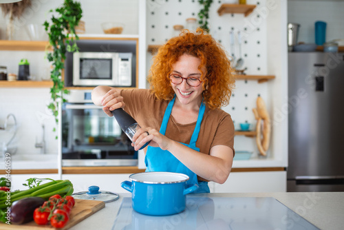 Cute young woman cooking and adding spice to meal, laughing and spending time in the kitchen