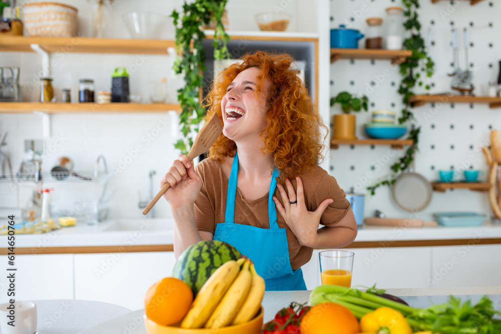 Funny beautiful woman singing into spatula, cooking in modern kitchen, holding spatula as microphone, dancing, listening to music, playful girl having fun with kitchenware, preparing food.