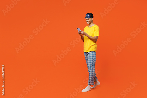 Full body side view young man wears pyjamas jam sleep eye mask rest relax at home hold in hand use mobile cell phone isolated on plain orange background studio portrait. Good mood night nap concept.