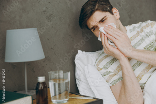 Young sick ill man with runny nose in casual clothes t-shirt pajama lying in bed sneeze hold napkin cough look at medicaments water spend time in bedroom home in hotel room wake up be in bad mood day. photo