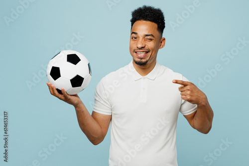 Young man fan wear basic t-shirt cheer up support football sport team hold in hand point index finger on soccer ball watch tv live stream wink blink eye isolated on plain pastel blue color background.