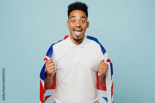 Young overjoyed exultant fun man fan wearing basic t-shirt cheer up support football sport team hold British flag do winner gesture watch tv live stream isolated on plain pastel blue color background.