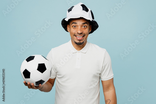 Young smiling fun happy man fan wear basic t-shirt hat looking camera cheer up support football sport team hold in hand soccer ball watch tv live stream isolated on plain pastel blue color background. © ViDi Studio