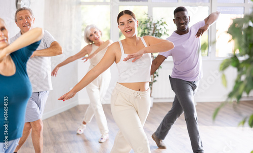 Young girl coach oversees mentoring group of elderly clients in fitness studio. Multiracial people group of different ages practice repeating dance movements