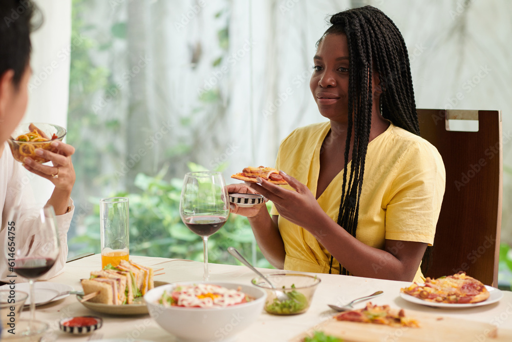 Smiling Black young woman enjoying slice of homemade pizza at dinner with friends