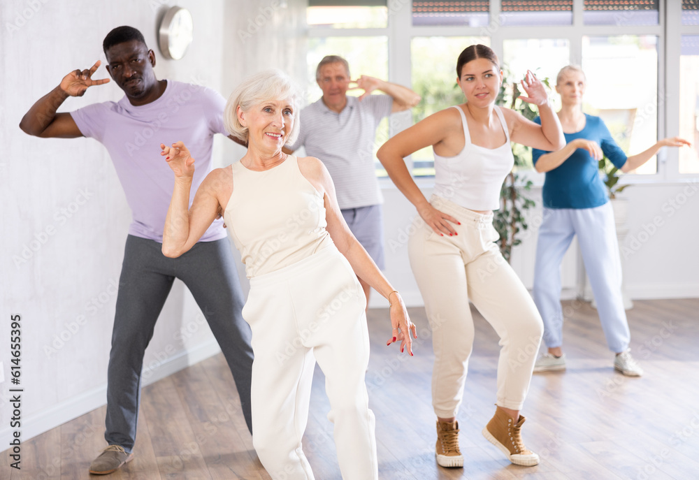 Fototapeta premium ..Young woman teacher teaches group of multiethnic mature active people dancing modern dance moves in studio