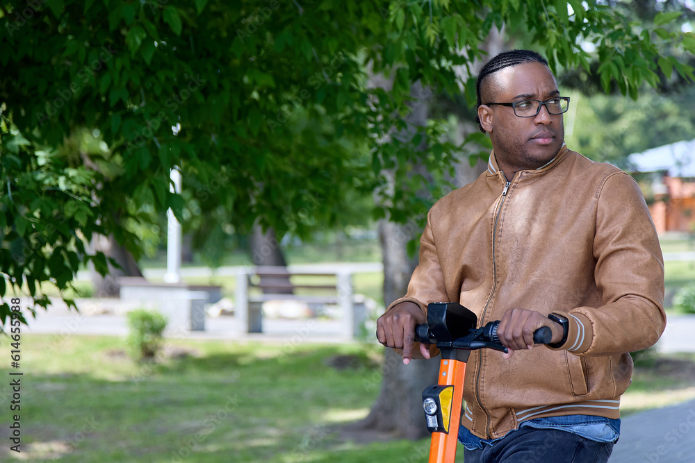 Dark-skinned guy in glasses and fashionable jacket walking along alley electric scooter looked to side. Against backdrop green foliage trees, portrait of stylish adult African American riding scooter