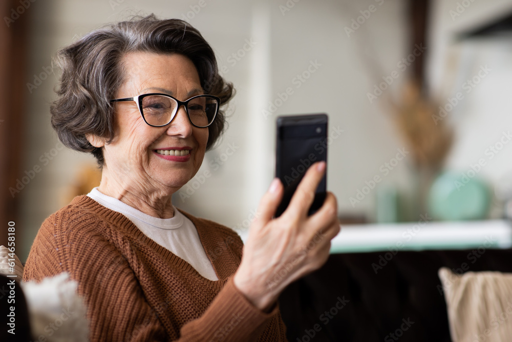 Happy mature middle aged woman enjoying using mobile apps, texting, sitting on sofa, smiling senior woman holding smart phone, looking at mobile phone screen, browsing social media at home Happy mature middle aged woman enjoying using mobile apps, texting, sitting on sofa, smiling senior woman holding smart phone, looking at mobile phone screen, browsing social media at home
