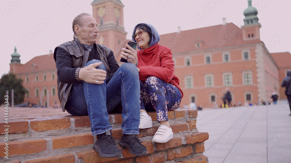 Elderly couple of tourists looking at their mobile phone are sitting in the historic center of an old European city and talking to each other. Palace Square, Warsaw Old Town