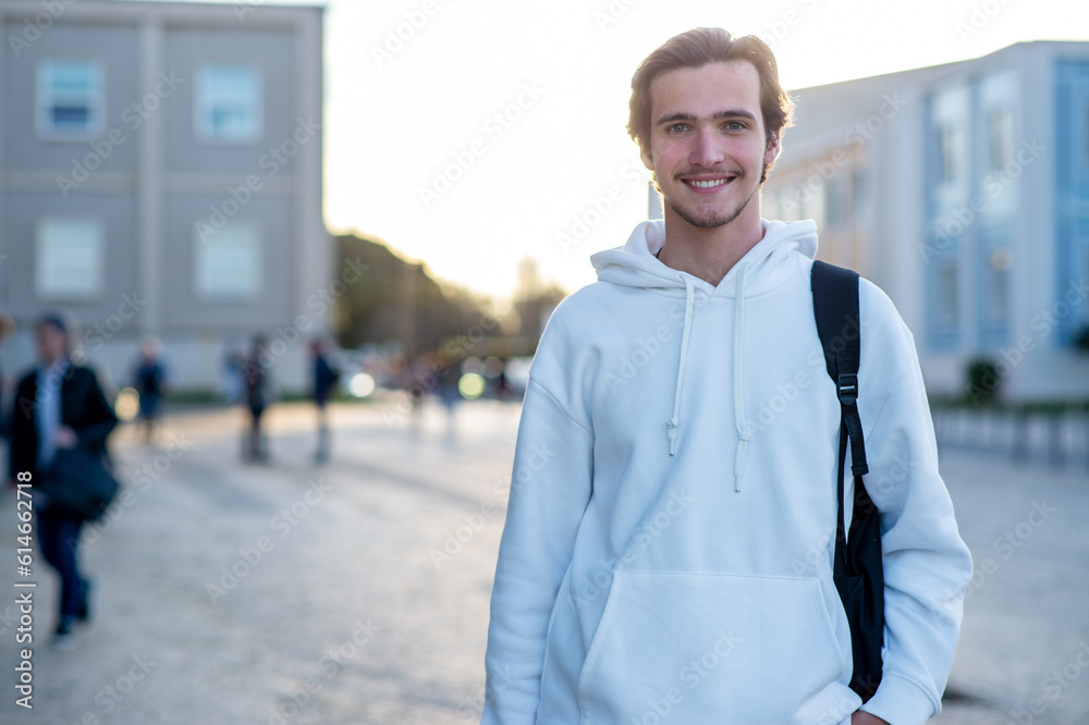 Happy man standing on a sidewalk near his faculty building with a backpack. Student smiling while standing on campus at his university. A cheerful young man with a backpack outdoors. Happy man standing on a sidewalk near his faculty building with a backpack. Student smiling while standing on campus at his university. A cheerful young man with a backpack outdoors.