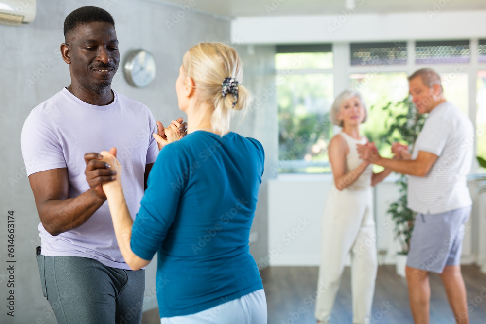 Fototapeta premium Happy smiling elderly woman enjoying impassioned merengue with african american partner in latin dance class. Social dancing concep