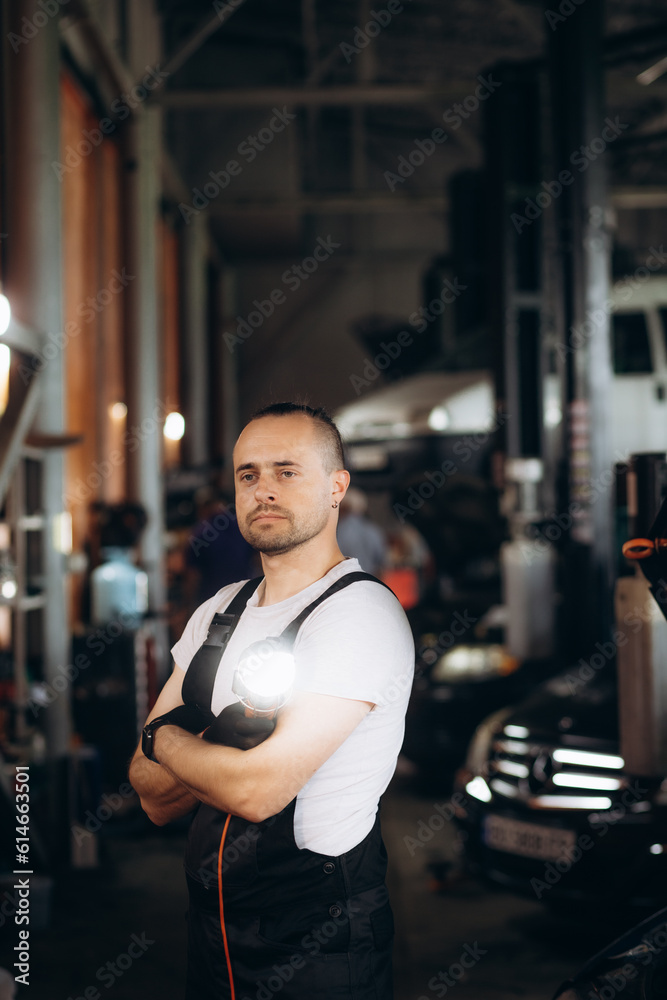 Handsome Car Mechanic is Posing in a Car Service. He Wears a Jeans Shirt and Safety Glasses. His Arms are Crossed. Specialist Looks at a Camera and Smiles