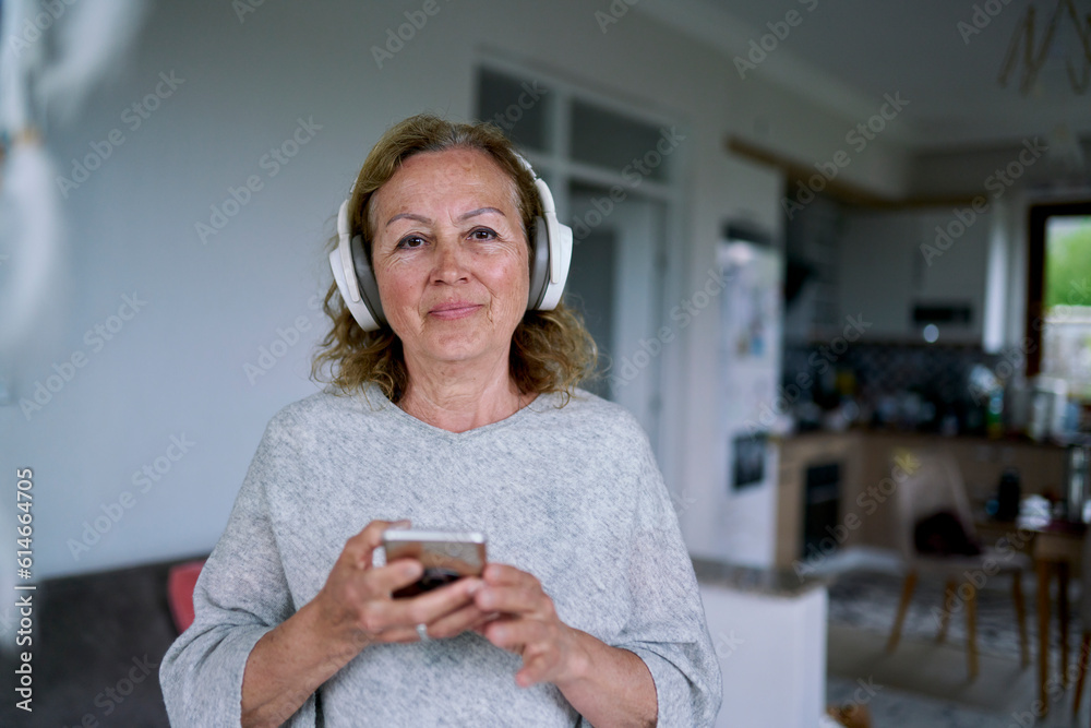 Senior woman wearing wireless headphones standing with smart phone at home Senior woman wearing wireless headphones standing with smart phone at home