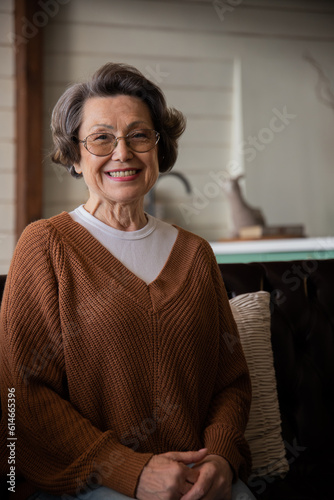 Happy old woman in glasses posing at home indoors, positive lonely senior pensioner woman sitting in living room portrait
