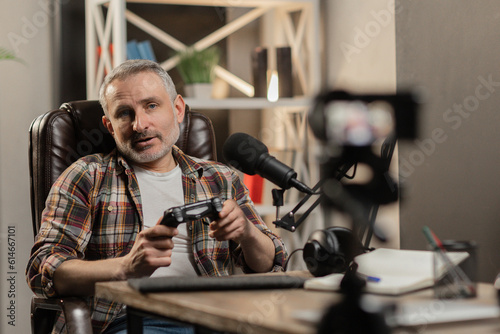 A gray-haired man in a plaid shirt sits in front of the camera and holds a gamepad in his hands. A male tech blogger is recording a video review of a new gamepad.