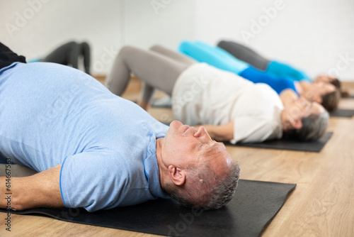 Diligent elderly man practicing pilates on gray mat in exercise room during pilates classes. Persons doing pilates in fitness hall