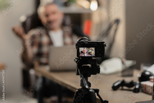 A man sits at a table in front of a laptop and records video on the camera. A male blogger is recording a new video using a small camera on a tripod.