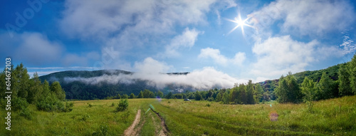 Panorama of the mountainous countryside near the forest. Morning fog melts under the rays of the bright summer sun. Dirt road invites for a morning walk