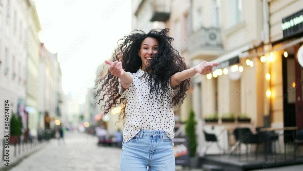 Beautiful young hispanic woman makes a beckoning gesture with one finger, inviting to approach, looks playfully flirting. Gorgeous girl waving hand to come with me on city street. Come here, follow me