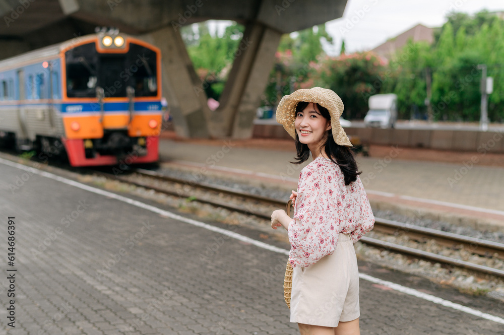 young asian woman traveler with weaving basket looking back happy smiling to a camera beside train coming background. Journey trip lifestyle, world travel explorer or Asia summer tourism concept.