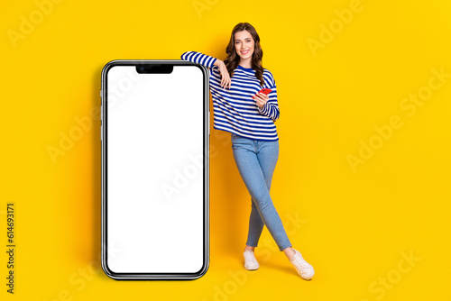 Full size photo of attractive young woman lean on big smartphone hold gadget wear trendy striped look isolated on yellow color background