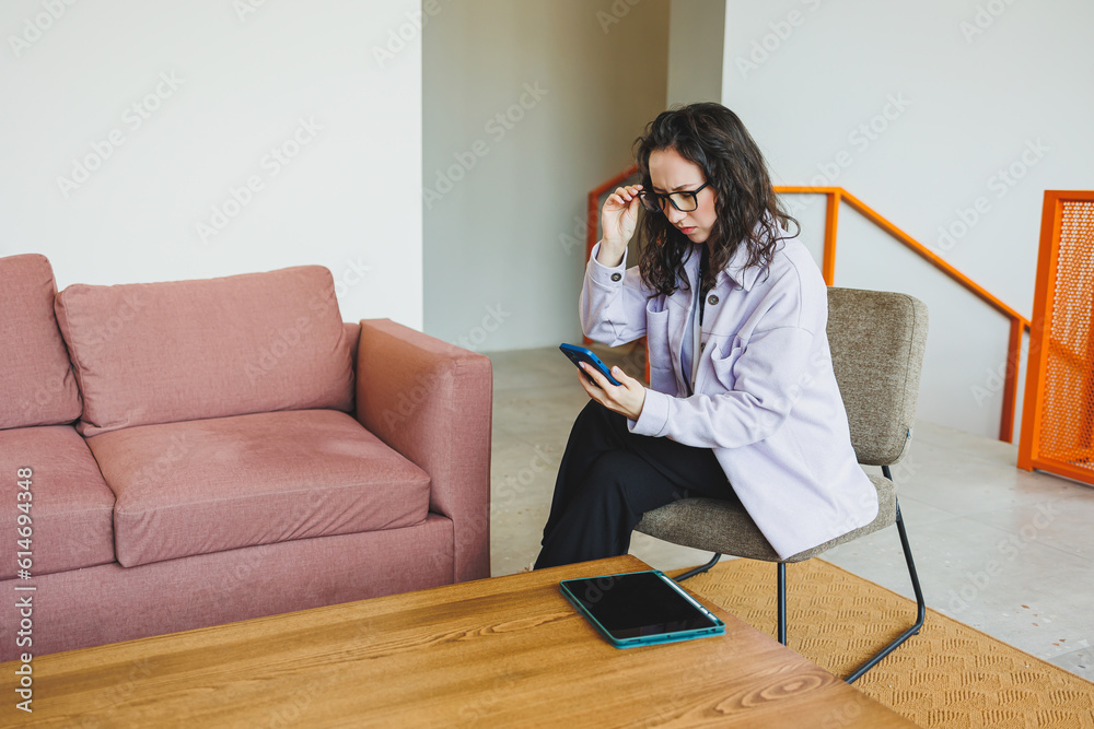 Emotional pretty woman in glasses in casual clothes sitting in comfortable chair in modern cafe at table with phone working online. Remote work Emotional pretty woman in glasses in casual clothes sitting in comfortable chair in modern cafe at table with phone working online. Remote work
