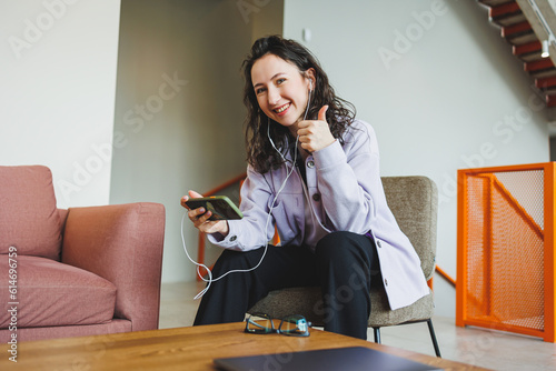 A stylish woman in glasses sitting in a modern cafe at a table with a phone works online. A woman is talking on the phone through headphones. Remote work. Work of a freelancer