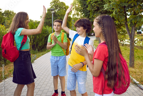 Happy little children boys and girls with backpacks saying goodbye after school or extracurricular activities applauding and high five to each other stand on path in summer park. Friendship, childhood