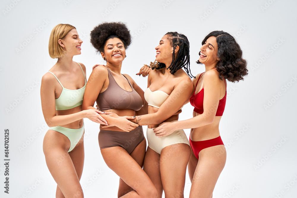 Cheerful multiethnic women in colorful lingerie hugging african american friend while standing and posing together isolated on grey, different body types and self-acceptance concept