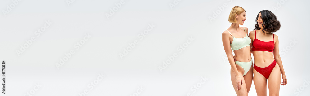 Cheerful multiracial woman in modern lingerie hugging blonde friend and standing isolated on grey, different body types and self-acceptance concept, multicultural models, banner Cheerful multiracial woman in modern lingerie hugging blonde friend and standing isolated on grey, different body types and self-acceptance concept, multicultural models, banner