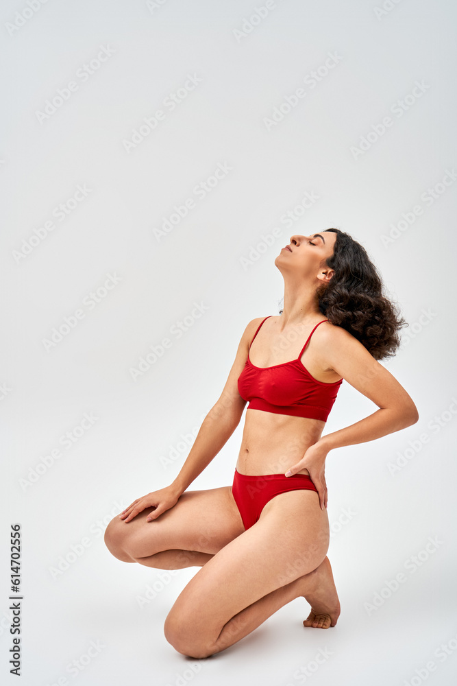 Side view of middle eastern brunette woman in modern red lingerie holding hand on hip and posing with closed eye on grey background, self-acceptance and body positive concept