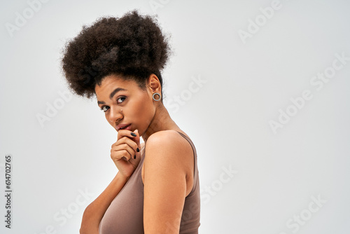 Portrait of confident african american woman in modern brown bra touching chin and looking at camera while standing isolated on grey, self-acceptance and body positive concept