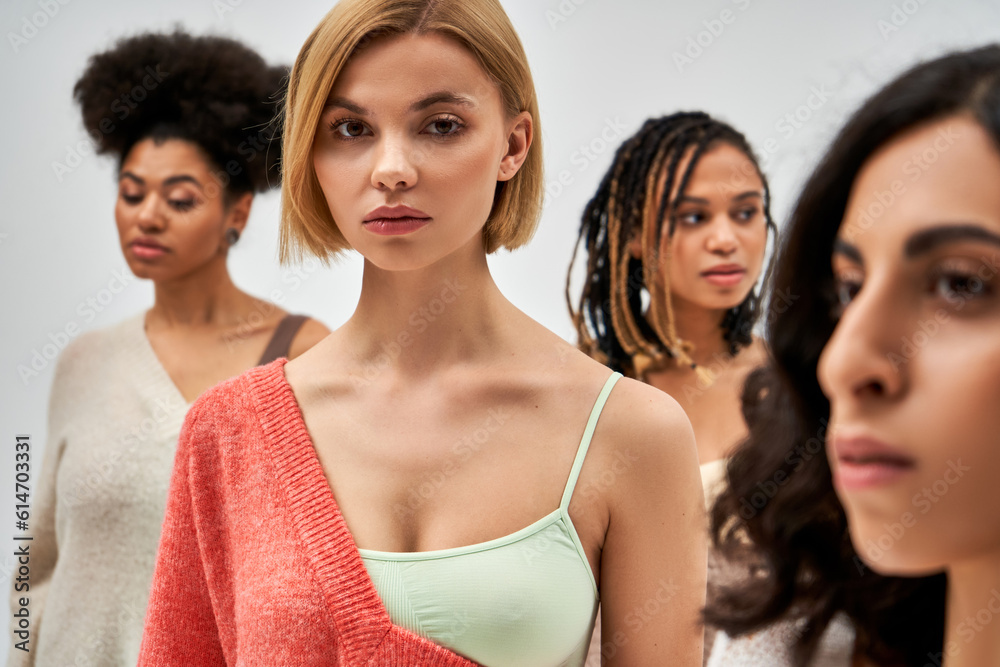 Portrait of young blonde woman in bra and wool jumper looking at camera while standing near blurred friends isolated on grey, different body types and self-acceptance, multicultural representation