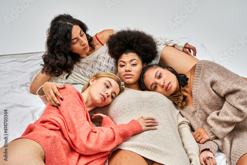 High angle view of multiethnic women in stylish warm jumpers looking at camera while lying on bed isolated on grey  different body types and self-acceptance  multicultural representation