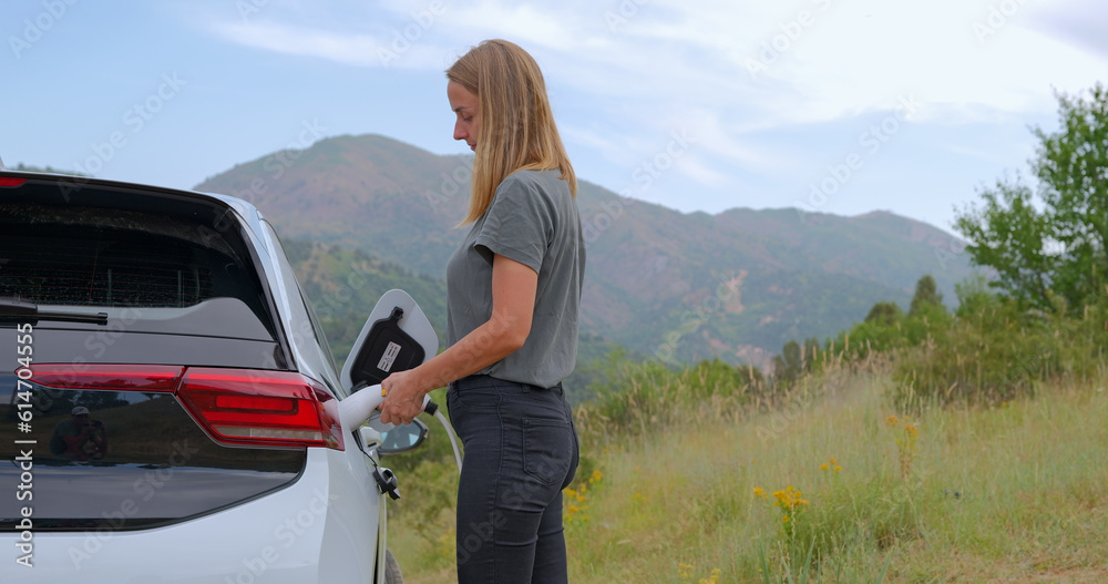 Young woman plugging charger in electric car, on mountains background Young woman plugging charger in electric car, on mountains background