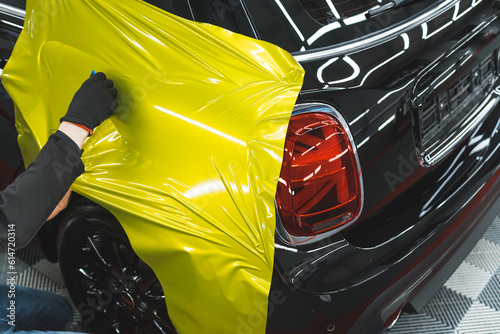 Unrecognizable car wrapper in black protective gloves using squeegee to straighten neon yellow vinyl film. High quality photo
